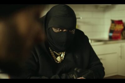 Masked person wearing gold chains and gloves at a kitchen table, pengar ar