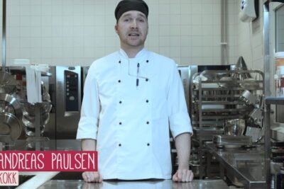 Chef in white coat and black cap standing in a stainless steel commercial kitchen, personer coliaki.