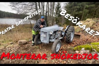 A man leaning on a grey vintage tractor near a forested lakeshore with autumn foliage, personliga asikter