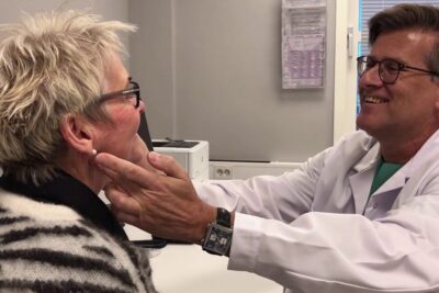 Smiling physician examines an older woman's jaw and ear during a Facial Consultation at plastisk kirurgi 2 Clinic – Expert Aesthetic Assessment in a medical office.