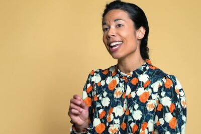 Smiling woman speaking in a floral dress against a warm yellow background illustrating positiivinen syrjinta.