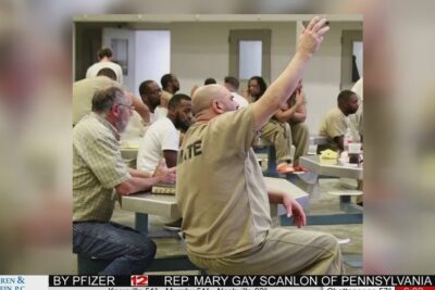 Inmates seated at tables in a prison dayroom with one man raising his hand during a group session, prison preventive 1.