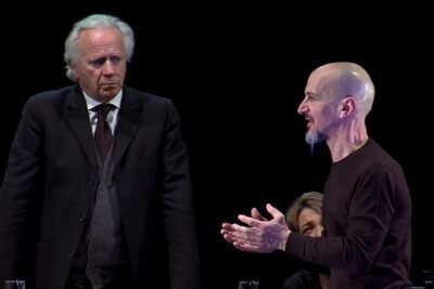 Two men on a dark stage engaged in intense discussion, evocative of a processo gesu