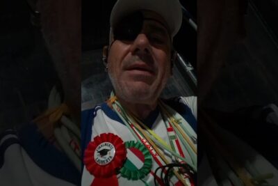 Close-up selfie of a man wearing a cap and eye patch with red and green rosettes and multiple lanyards, processo pace 1.