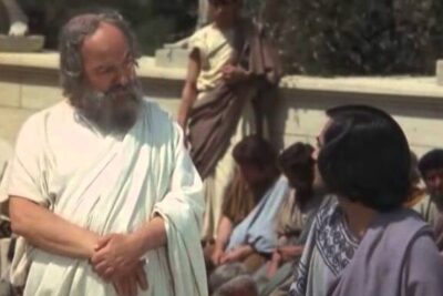 Bearded philosopher in a white toga speaking to a younger man during a processo socrate style trial scene
