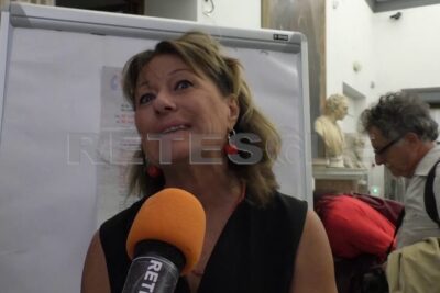 Person speaking into an orange microphone in front of a flipchart at an indoor event about promuovere luguaglianza.