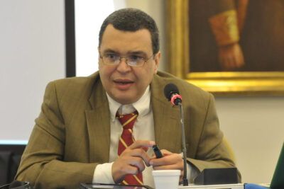 Conference speaker in a brown suit speaking into a microphone at a table with a cup and laptop, suitable for propiedad privada topics.