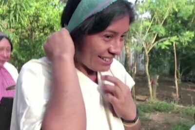 Smiling woman adjusting a headband outdoors with companions and a child in a wooded rural setting, pueblos indigenas 1.