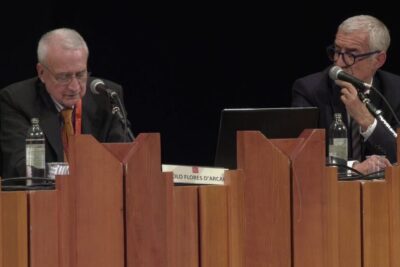 Two panelists seated behind a wooden podium speaking into microphones illustrating questione delleutanasia