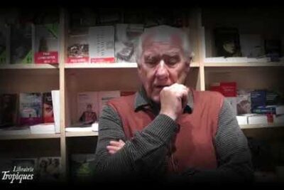 Elderly man seated in front of bookshelves with hand on chin, pondering realisme nietzsche