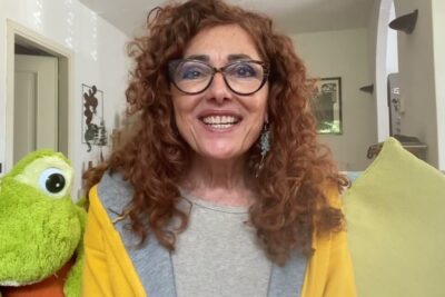 Smiling woman with curly red hair and glasses beside a green frog plush on a couch in a bright living room, reflecting realta oggettiva.