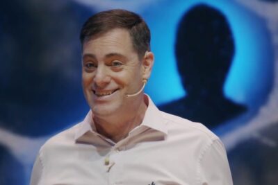 Smiling presenter wearing a headset microphone in front of a vivid blue circular backdrop and silhouetted profile, redes sociales 17