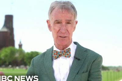 Man in green blazer and colorful bow tie speaking on location near a monument during a news segment about ways to reduce plastic.
