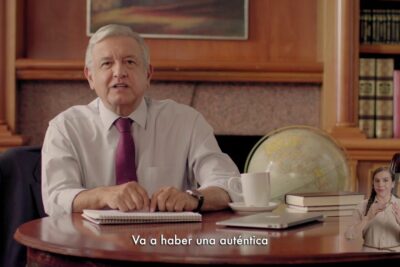 Man seated at a wooden desk delivering a speech about reforma educacional 1 with subtitles and a sign language interpreter