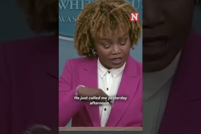 Speaker at a press podium in a pink blazer delivering remarks about regeringens politik.