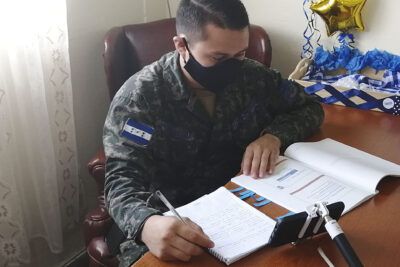 Person in camouflage uniform wearing a mask writing in a notebook while viewing a smartphone on a tripod, relevante 2.