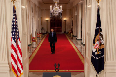 A man in a dark suit walks down a long red carpeted ceremonial corridor flanked by flags and columns, relevante 8.