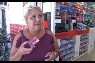 Woman holding a small boxed product at a convenience store counter, renda basica 1