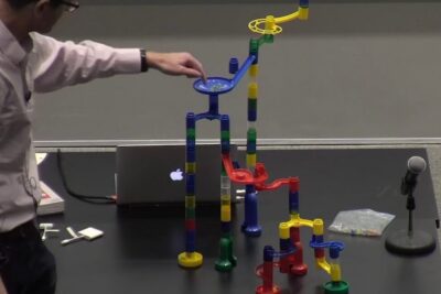 Person adjusting a tall colorful marble run toy on a lecture table with laptop and microphone, reproductive cloning 1