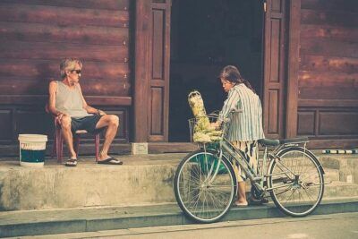 Woman with bicycle loaded with produce speaking to a seated man on a stoop, illustrating retfardig handel.