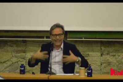 A speaker gestures behind a table during a lecture with bottles, notes and a projection screen visible, rifiuto bambino 1