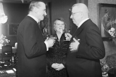 Three formally dressed people in a vintage black-and-white interior, holding glasses and conversing with subtle ristiriidat.
