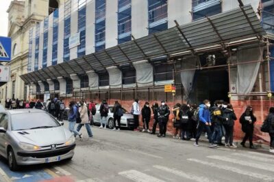 Students queuing on a city sidewalk outside a scaffolded school during a ritorno classe morning