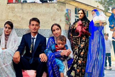 Seated couple in formal attire surrounded by family and children in vibrant traditional clothing at an outdoor gathering featuring rosta for