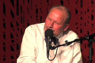 Older male singer in a white shirt and bow tie performing into a microphone, roykeloven norge