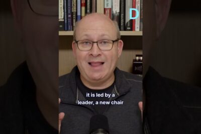 Middle-aged man speaking to camera in front of a bookshelf with a microphone visible, sahkoinen aanestys 1