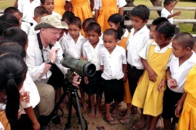 Schoolchildren in white shirts and yellow uniforms gathered around an instructor demonstrating a spotting scope, samfund 8.