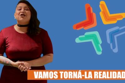 Presenter in a maroon dress stands against a blue background with a multicolored cross-style logo and an orange banner with white text, saude universal 1.