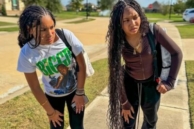 Two teenage girls standing on a suburban sidewalk with backpacks and focused expressions, sch ler 19