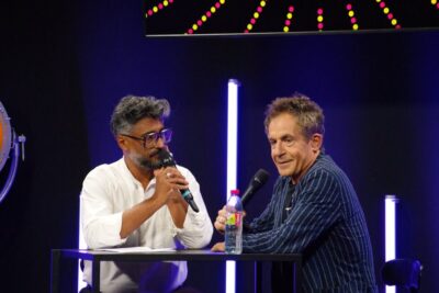 Two presenters holding microphones at a small studio table under neon lights during a science eclairee talk