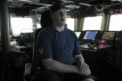 Crew member seated on a ship's bridge amid navigation screens and controls, sen tehtavana.