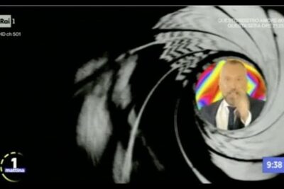 Gun-barrel style broadcast graphic with a suited presenter portrait framed by a circular target evoking sicurezza nazionale themes.