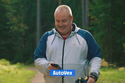 Middle-aged man smiling on a forest path holding a phone and a leash handle, siksi tarkeaa.