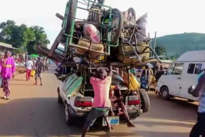 An overloaded car stacked with chairs, carts and bags moving through a crowded street market, simma strommen