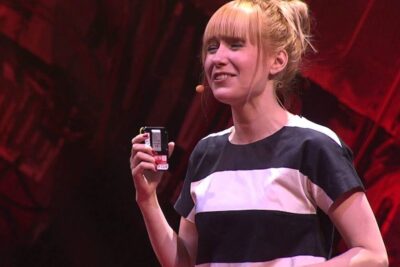 Woman speaker onstage holding a small electronic device while wearing a headset, illustrating a slike programmer presentation
