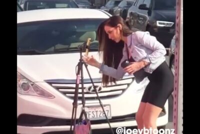 Woman in a light jacket adjusting a smartphone on a tripod beside a parked car on a busy street, social media 28.