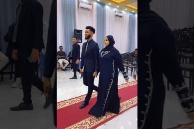 A bride in a navy embroidered dress and hijab and a groom in a dark suit walking hand in hand down a red carpet aisle, solmia avioliitto 1.