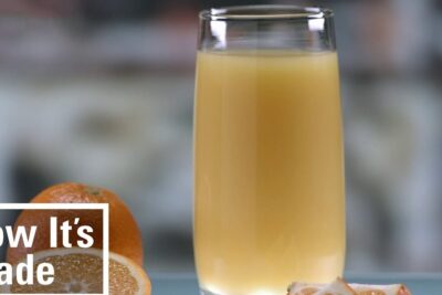 Tall glass of succo darancia 3 beside whole and sliced oranges on a countertop.