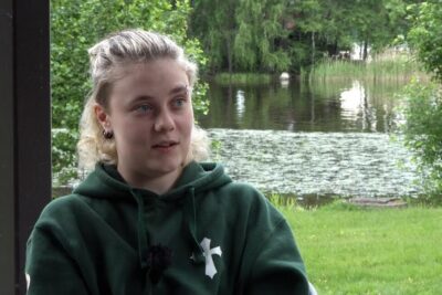 Person in a green hoodie seated outdoors by a pond, suitable illustration for sukupuolta oleville