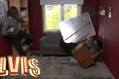 Two people tumble as furniture and a reflective panel collapse in a compact red-walled living room, tid for a chaotic scene.