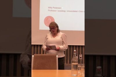 Speaker standing at a lectern presenting slides in a lecture hall about tidlig forebygging