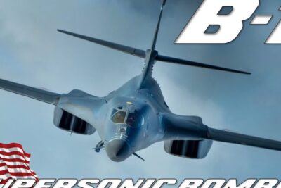 Front-facing B-1 supersonic bomber flying toward the camera with an American flag and bold overlay text, tilbagekaldelse embedet