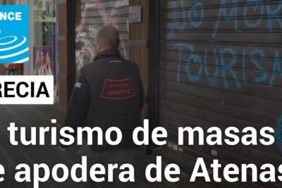 Graffiti reading 'No more tourism' on a closed Athens storefront with a passerby and a news headline — turismo masas 1