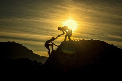 Silhouetted pair on rocky terrain at sunset with one person helping another climb, symbolizing tyottomyydesta ja sen vastustamisesta tyoton vai tyollistetty mielipiteita.