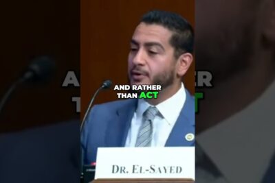 Panel speaker in a suit addressing a microphone at a formal hearing, illustrating universell helseforsikring debate.