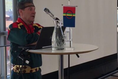 Person in traditional-style clothing at a round table with laptop, glass pitcher and small multicolor flag, urfolks rettigheter 1